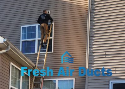Man on ladder cleaning exterior vent of house in Owings Mills, MD; "Fresh Air Ducts" logo and Air Ducts Cleaning text overlay at the bottom.