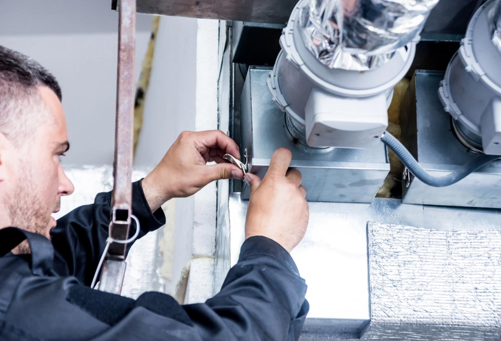 A person in work clothes adjusts air ducts and wiring in an HVAC system.