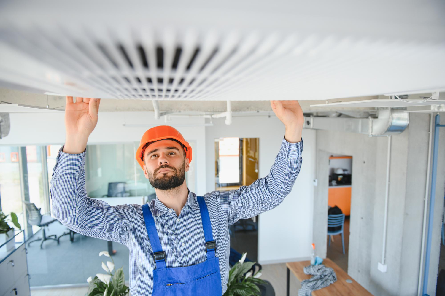 Man in a hard hat and overalls fixing or inspecting a ceiling air vent in a modern office.