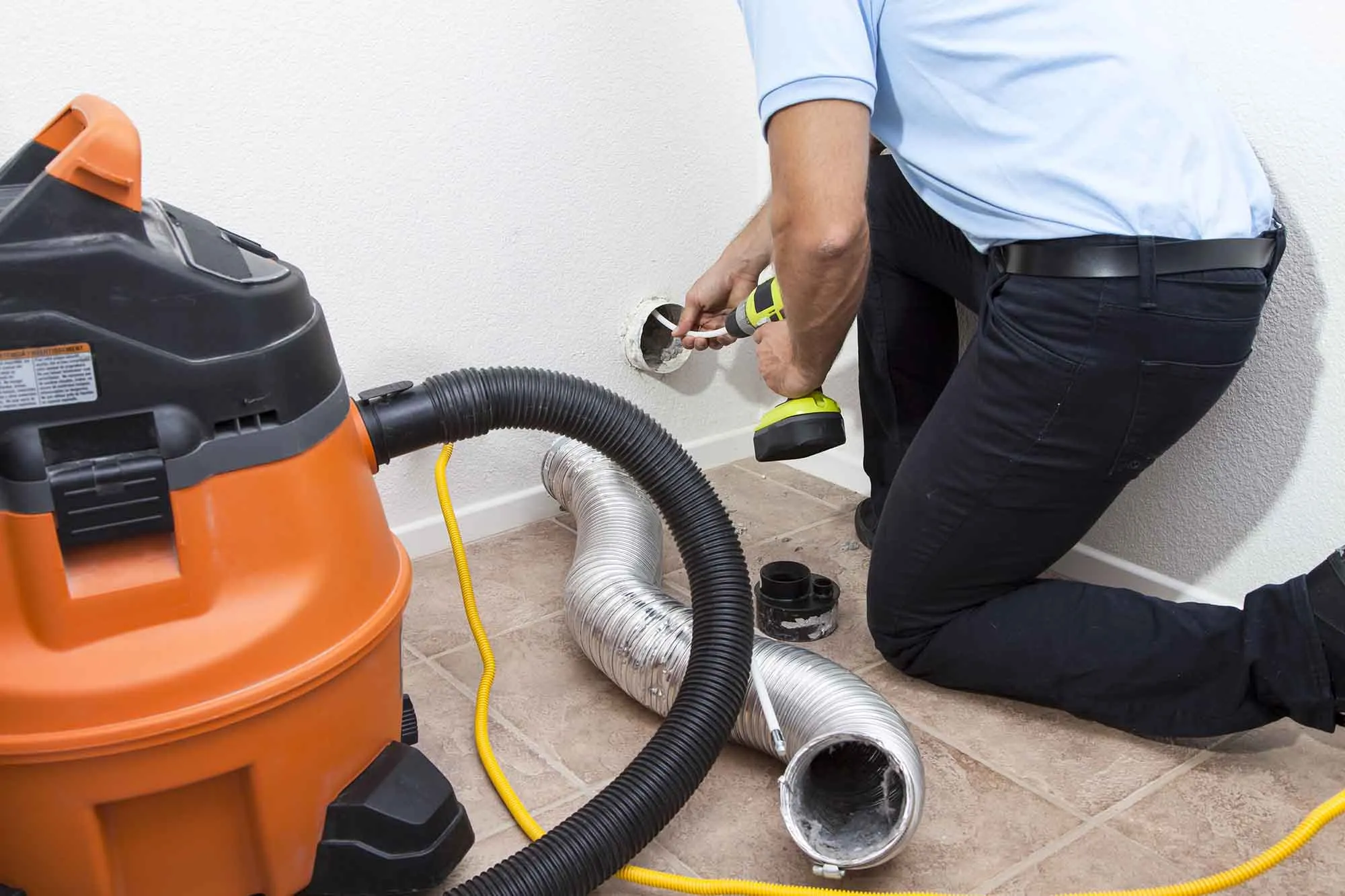 dryer-vent-hero A person cleaning a dryer vent with a vacuum and power tool next to a flexible duct on a tiled floor.