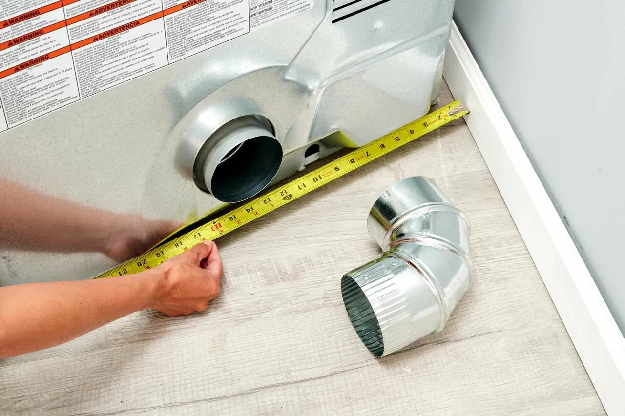 dryer-vent-install-1 A person measures behind a dryer vent with a tape measure; a metal duct elbow sits nearby on the floor.