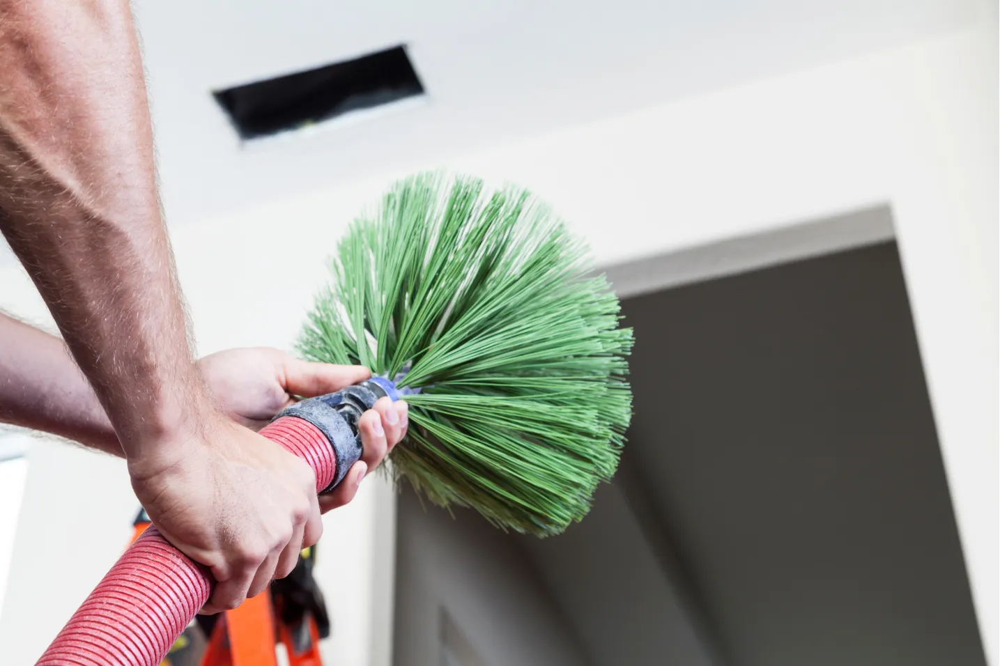 Person holding a brush tool, cleaning or inspecting an air duct vent on a ceiling indoors.