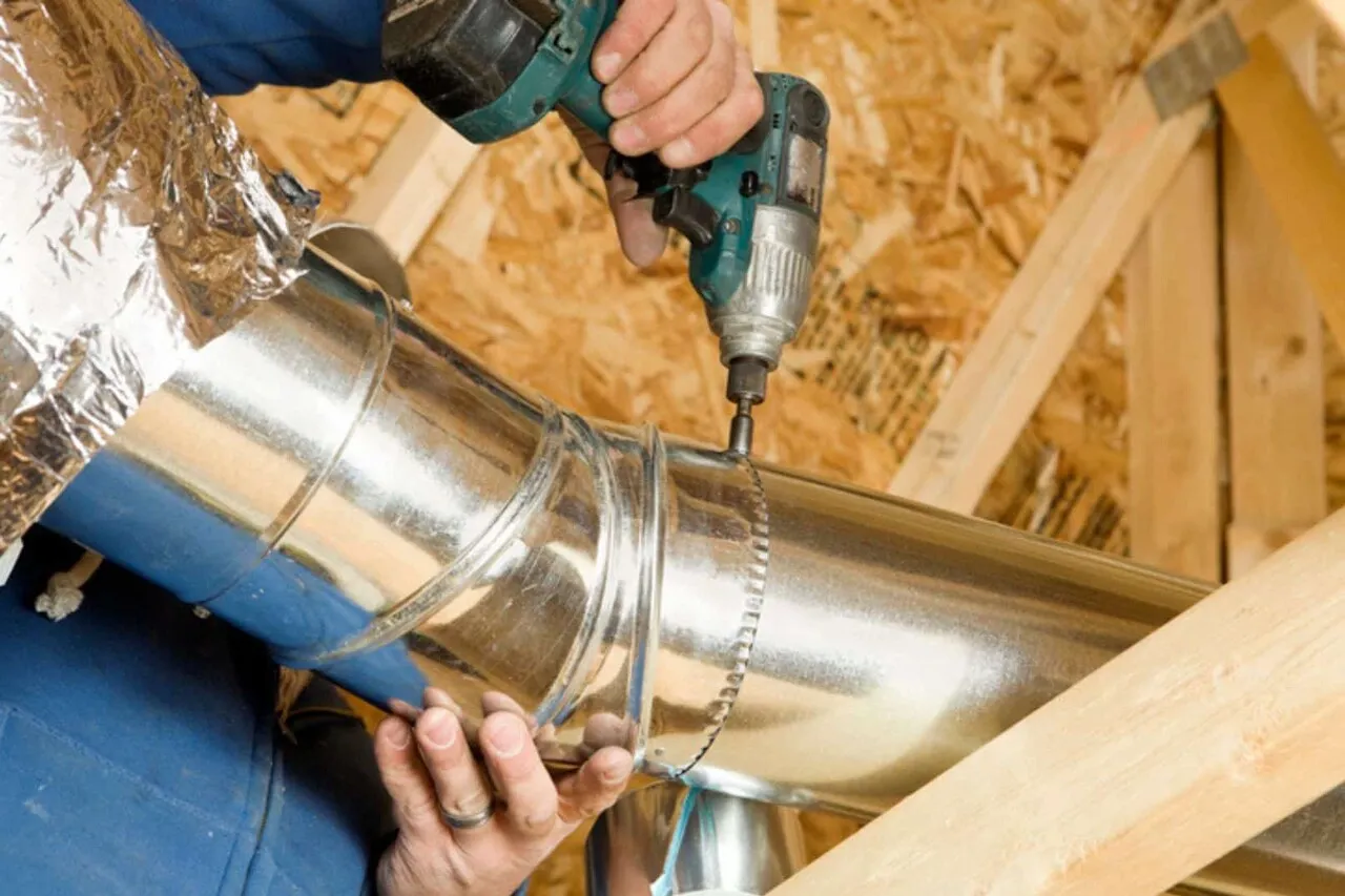 duct-install-1 Person using a power drill to install a metal air duct in a wooden attic or ceiling space.