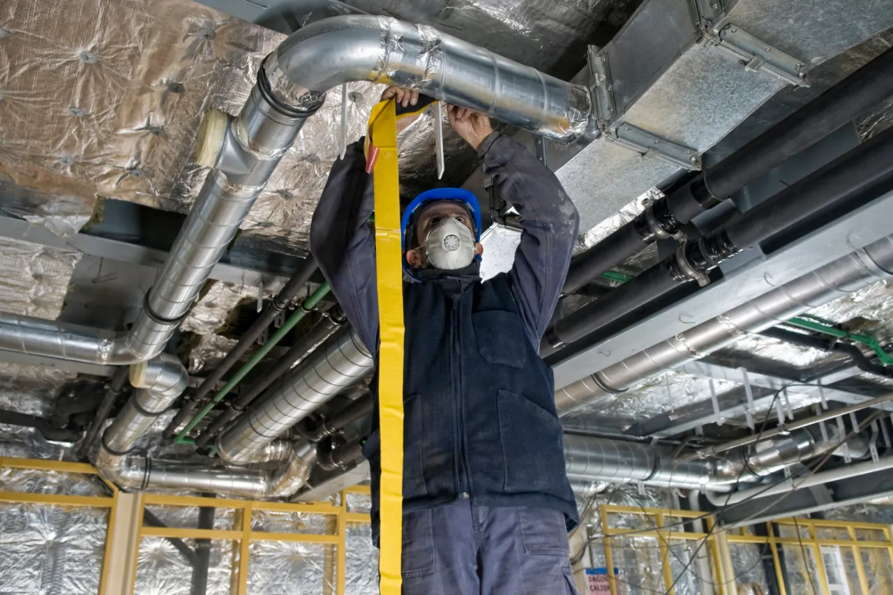 Worker wearing safety gear measures industrial ductwork on a ceiling with a yellow tape measure.