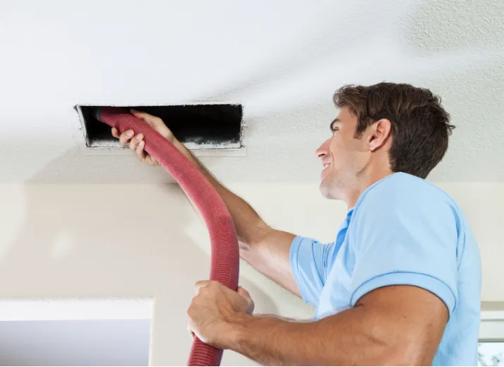 hvac-clean-1 Man cleaning an air duct on the ceiling with a large vacuum hose, wearing a light blue shirt.
