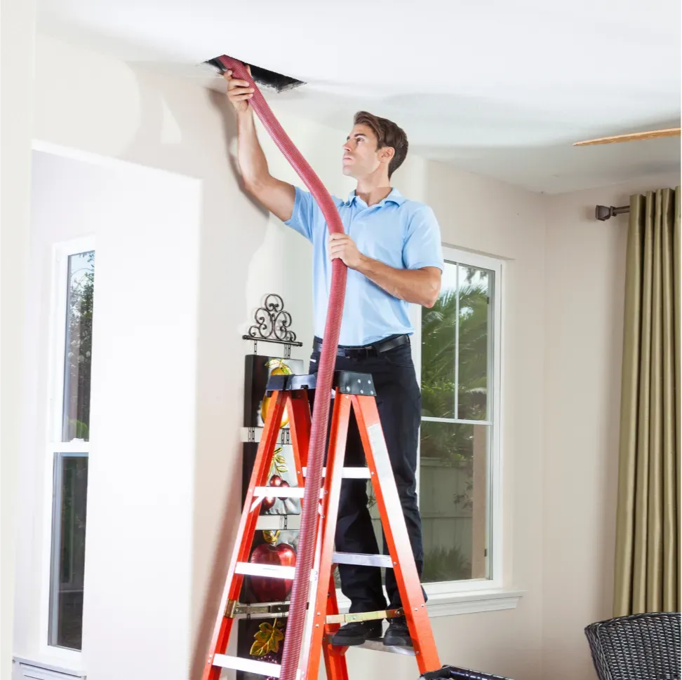 Duct Disinfectant: Man on ladder cleaning ceiling air vent with a vacuum hose in a bright living room.