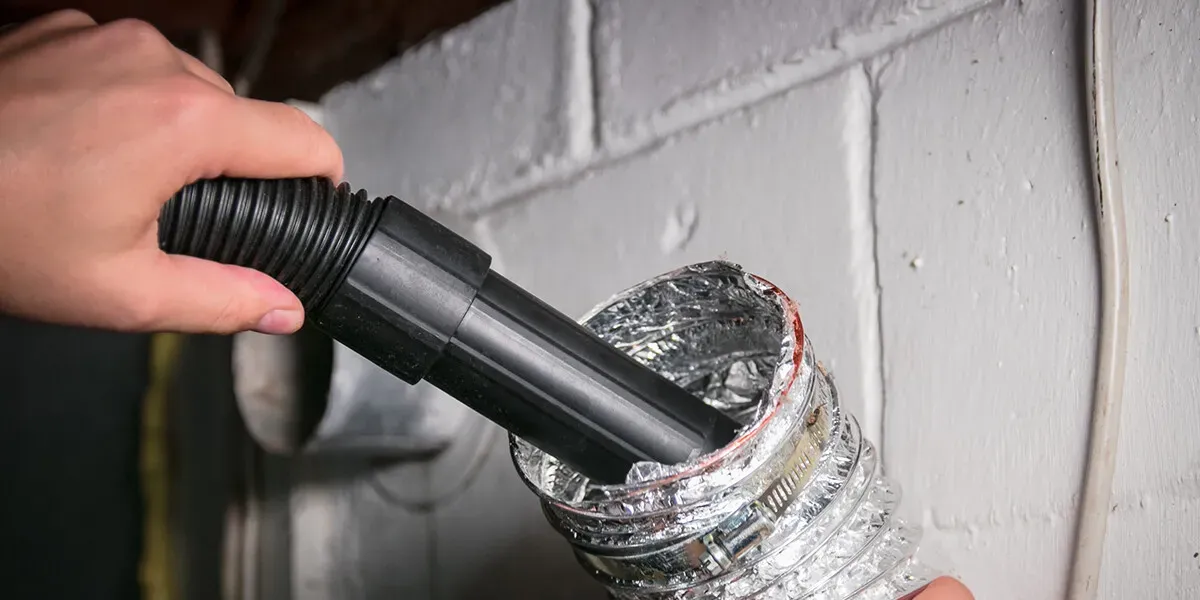 A person using a vacuum hose to clean inside a flexible dryer vent duct attached to a wall.