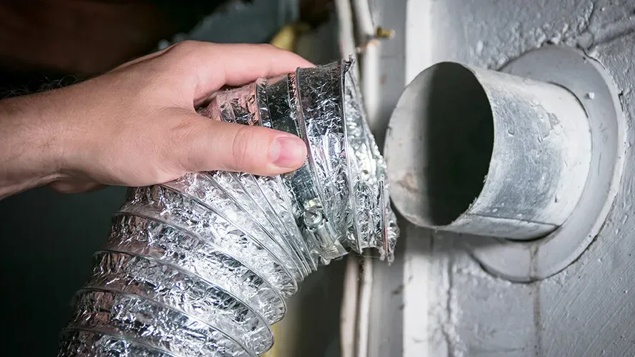 A hand connects a flexible metal dryer vent hose to a round wall duct opening.