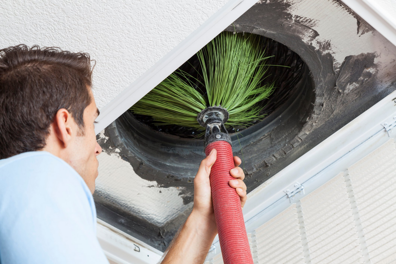 Man cleaning air duct with a brush attached to a vacuum hose, removing dust from ceiling vent.