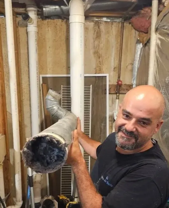 A man holds a dirty air duct, smiling in a room with exposed pipes and insulation.
