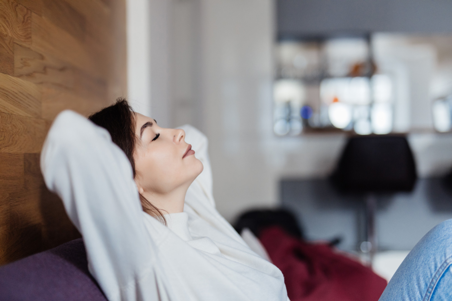 Woman relaxing on a couch with eyes closed and hands behind her head in a cozy indoor setting.